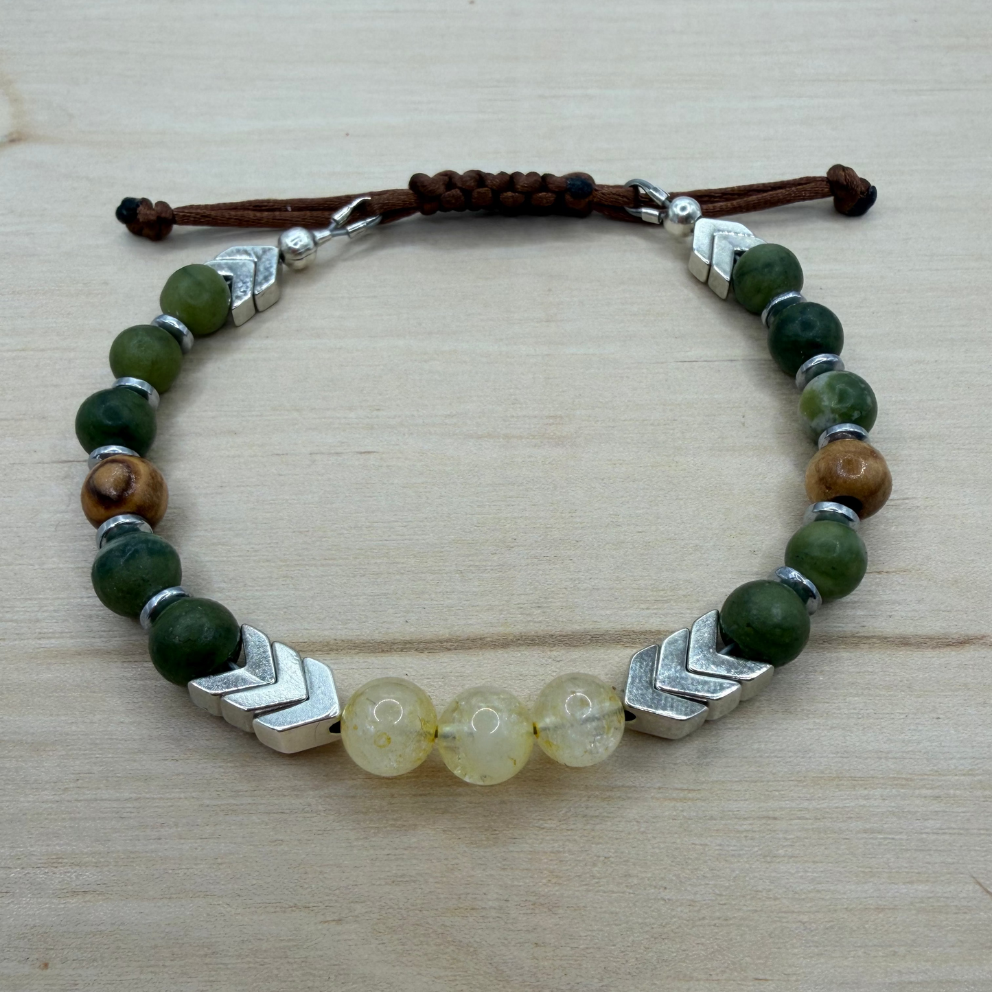 Bracelet with green beads, silver accents, and a brown cord on a wooden surface