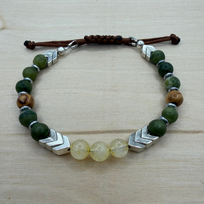 Bracelet with green beads, silver accents, and a brown cord on a wooden surface